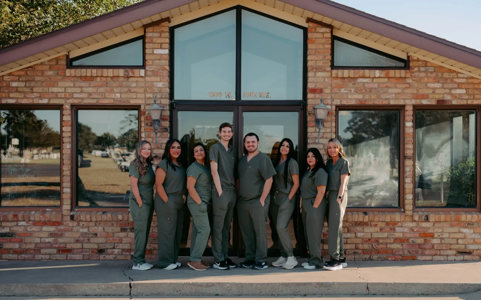 A team of eight staff members in green scrubs stands smiling in front of a brick building.