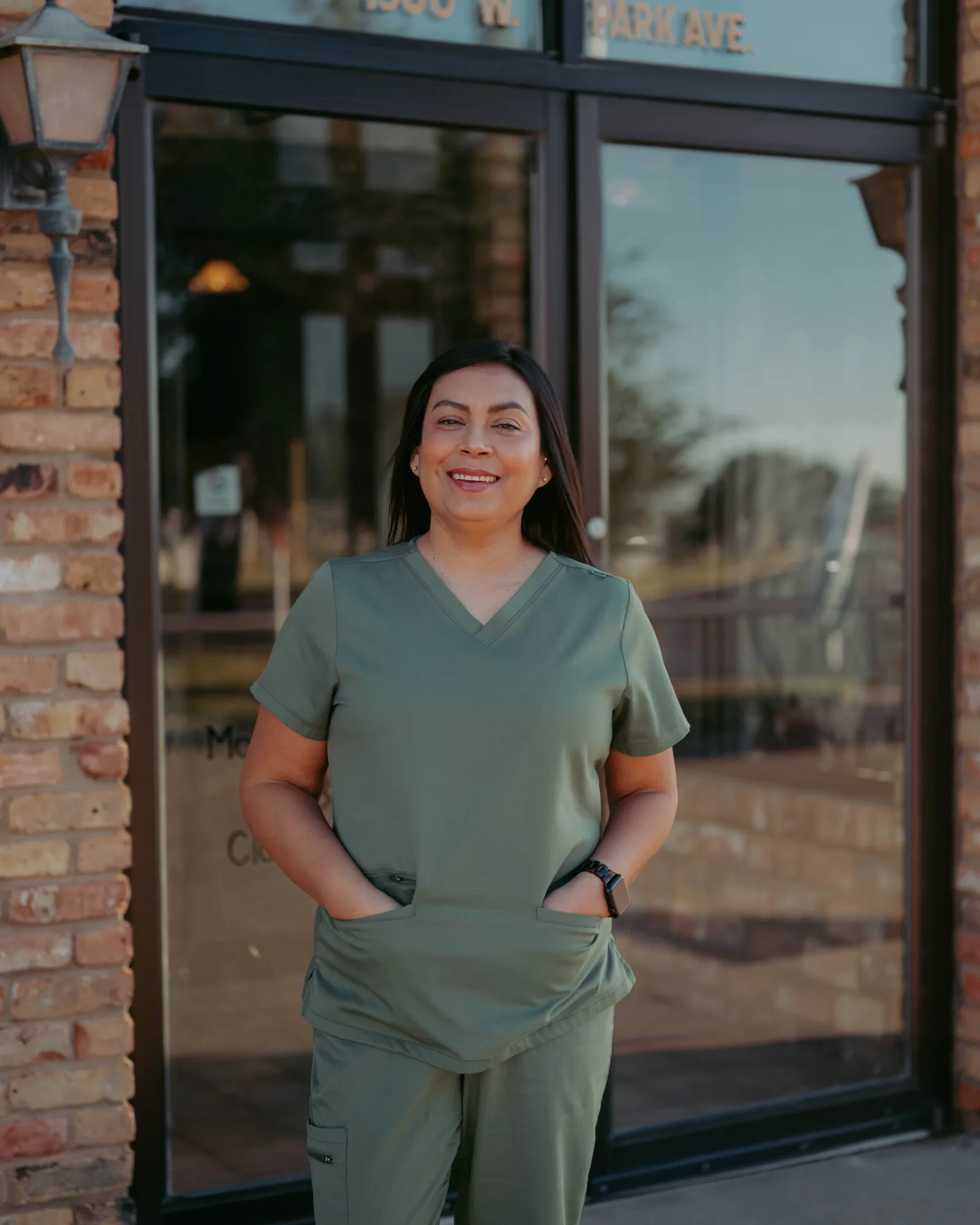 Smiling woman in green scrubs with hands in pockets.